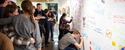 A crowd of students stand and watch three other students write on a large white wall. The wall holds writings from other students in a variety of colours.
