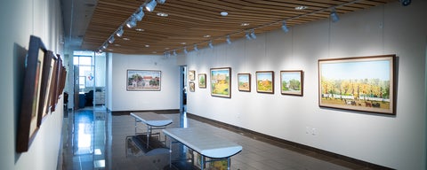A view of the gallery space, with three white walls, forming a long rectangular room. Paintings of landscapes hang on the white walls. Two benches sit in the middle.