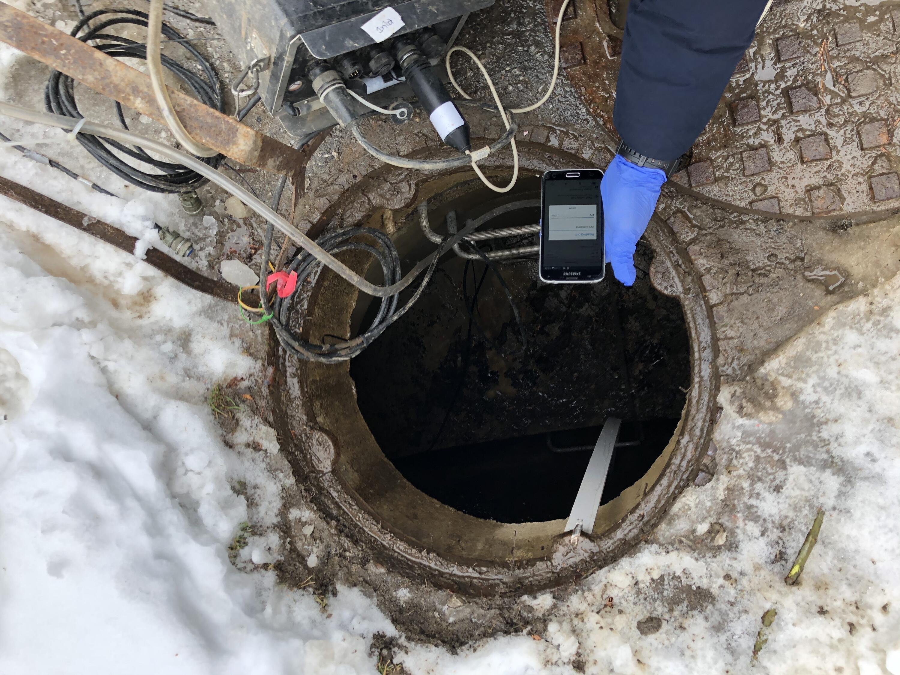 person's hand holding a cell phone over a maintenance hole to collect data from logger