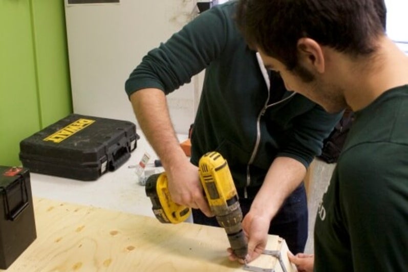 One student drilling hinge bracket to attach two pieces of wood, while another student holds on of the pieces