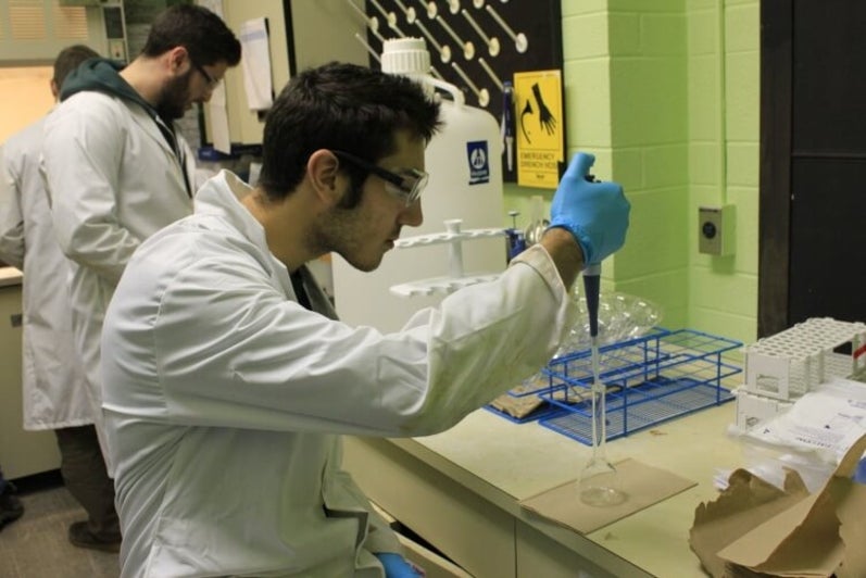 Student in lab coat sitting at bench and pipetting liquid into test tube
