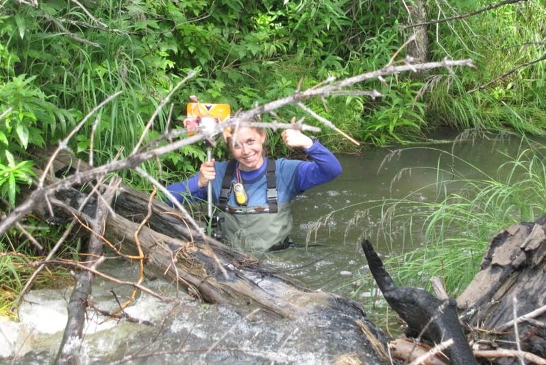 Female student in hip waders standing in waist deep water holding a metal rod with a yellow plate on the end