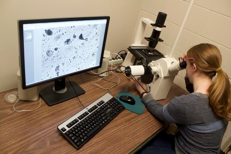Female student looks through microscope type maching with grey shapes showing on computer screen beside her