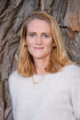 Deirdre Laframboise: middle-aged woman with blonde shoulder length hair standing in front of a large tree trunk wearing a beige sweater