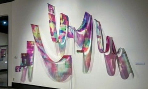 Artwork of a strip of vibrant transparent images that is looped over rods on a wall.