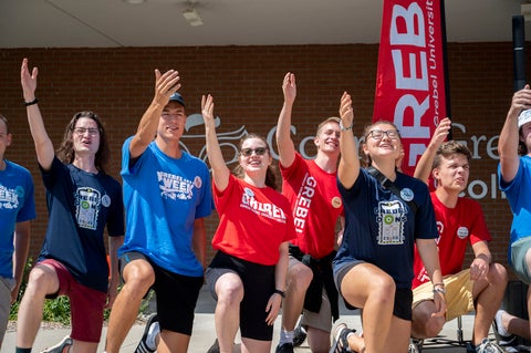 A group of upper year students in red and blue t-shirts perform an enthusiastic welcome on move-in day. They are all kneeling, with their arms raised, singing "We love you Grebel"