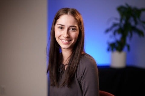 Milena Gojsevic, a Biochemistry student, is wearing a grey shirt and is smiling. There are white walls and a blue light in the background.
