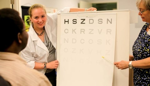 Client viewing Pelli-Robson contrast sensitivity chart while optometrist points to a letter and an intern observes.