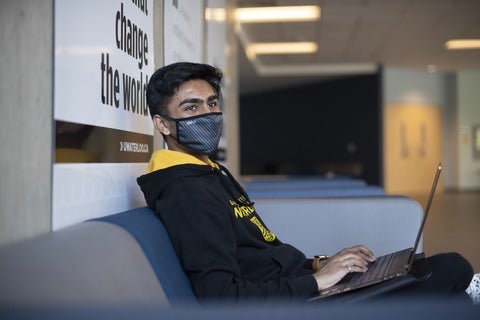 A University of Waterloo co-op student wearing a face mask and sitting indoors on a couch and using a laptop.