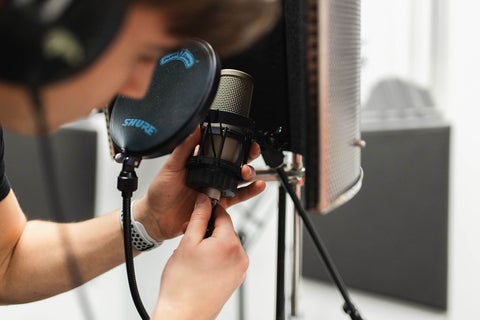 A student wearing headphones is plugging in an XLR cable to a microphone in the sound studio.